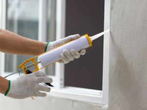 A close up of a person's hand smoothing a white caulk into a crack around a window, preventing drafts and increasing energy efficienc