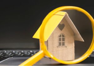This is an image of a small wooden house sitting on a laptop keyboard with a yellow magnifying glass in front of the house.