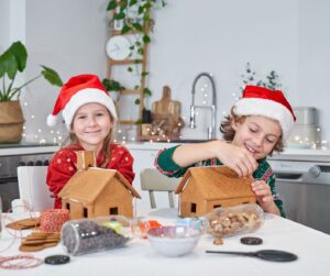 This is an image of a boy and a girls wearing Santa hats, sitting at a table decorating gingerbread houses.