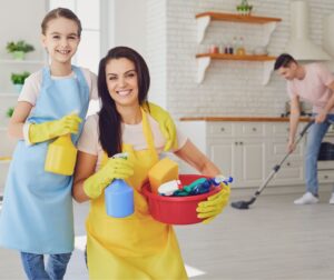 This is an image of a mother and daughter holding cleaning supplies with a dad in the background vacuuming.
