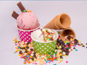 This is an image of 2 cups of ice cream, 3 empty cones and candy and sprinkles scattered in front of them.
