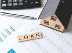 This is an image of the word loan spelled out in scrabble tiles, a wooden house on top of some budget paperwork.